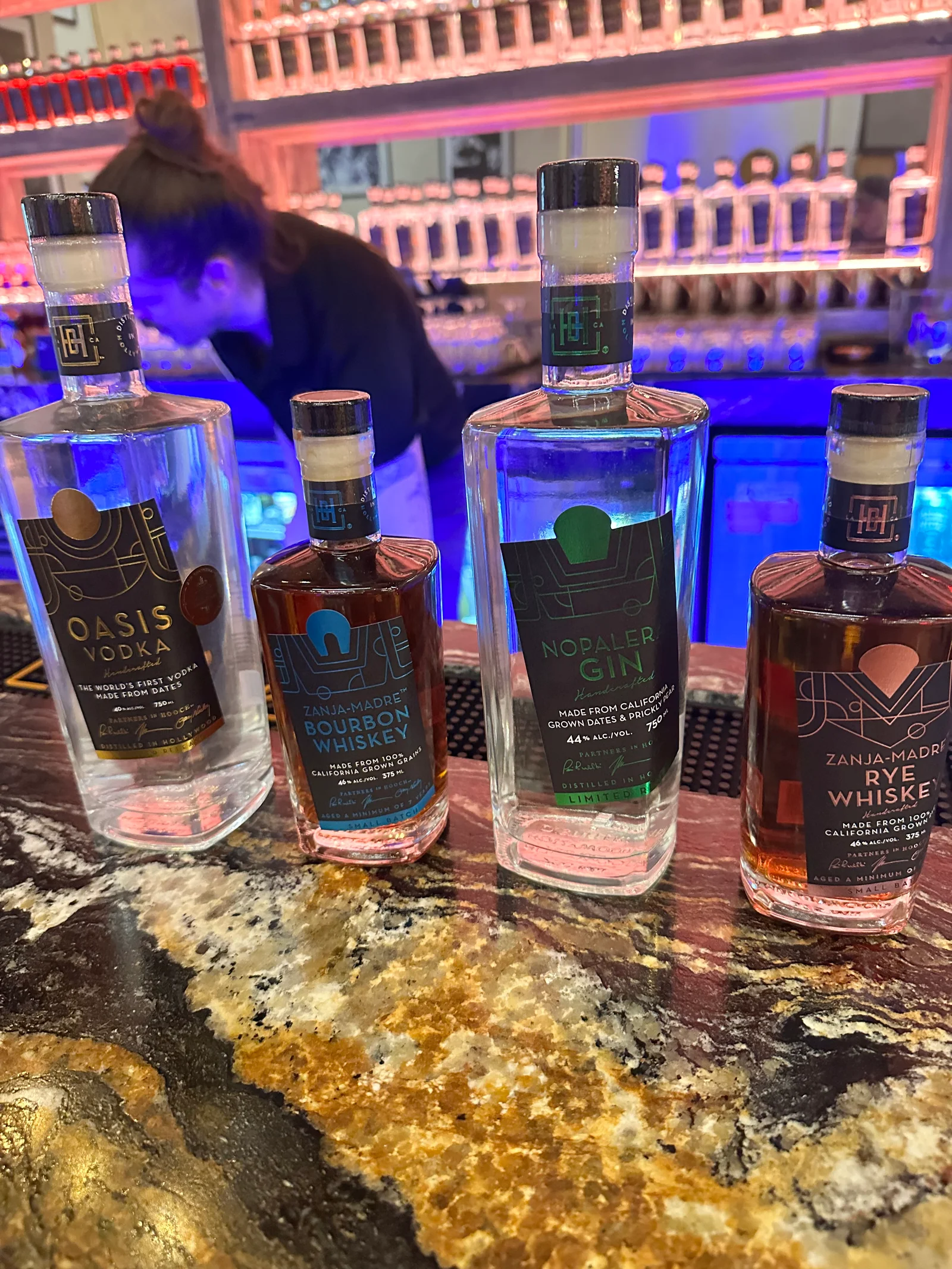 Four Hollywood Distillery bottles lined up on a gold and black granite bar: Oasis Vodka, Zanja-Madre Bourbon, Nopalera Gin and Zanja-Madre Rye Whiskey, with a bartender working in front of a backlit wall of bottles behind them