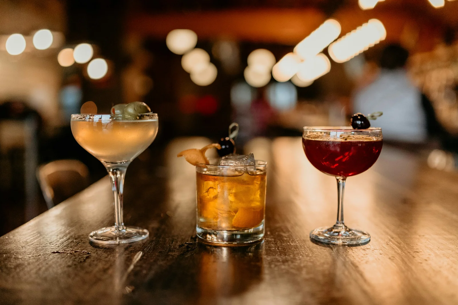 Three cocktails lined up on a wooden bar counter in a warmly lit bar: a pale drink with olives, an amber drink on ice with orange peel, and a deep red drink with a cherry
