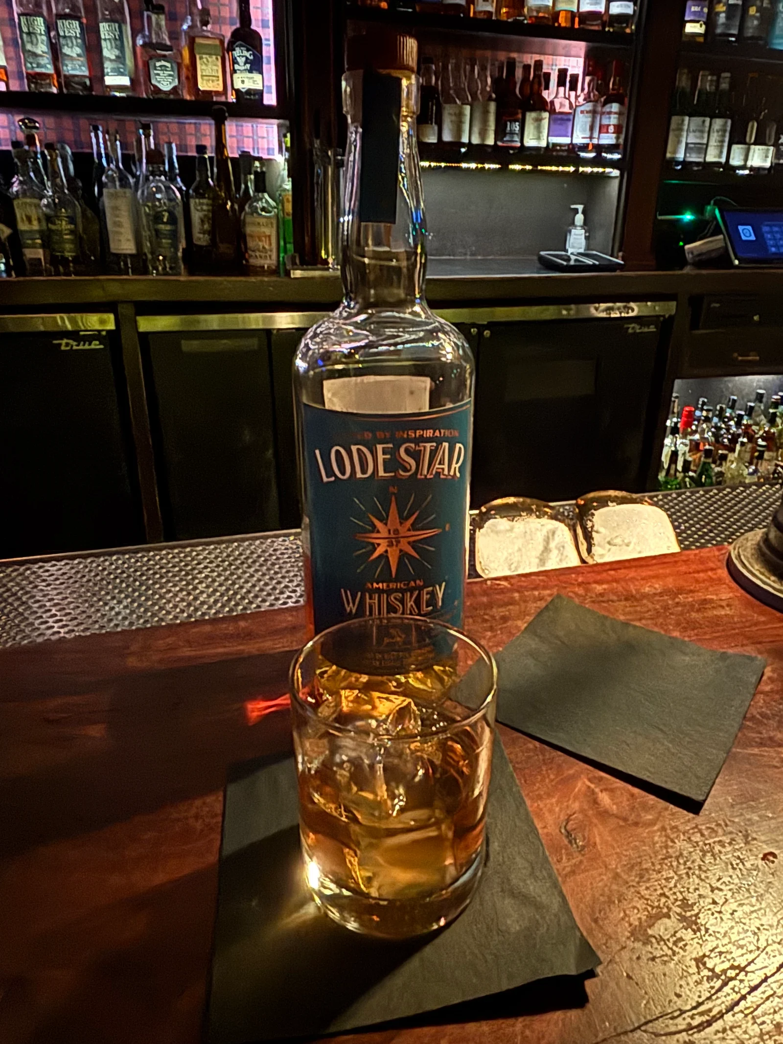 A bottle of Lodestar American Whiskey with its blue compass label beside a rocks glass of whiskey over ice on black napkins, on a worn wooden bar with backlit shelves of whiskey behind it